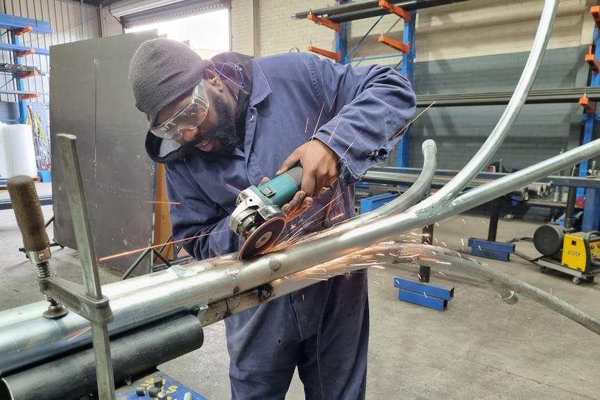 A man with dark skin and a black beard is dressed in blue overalls and uses a grinder on a metal pole positioned in a workshop