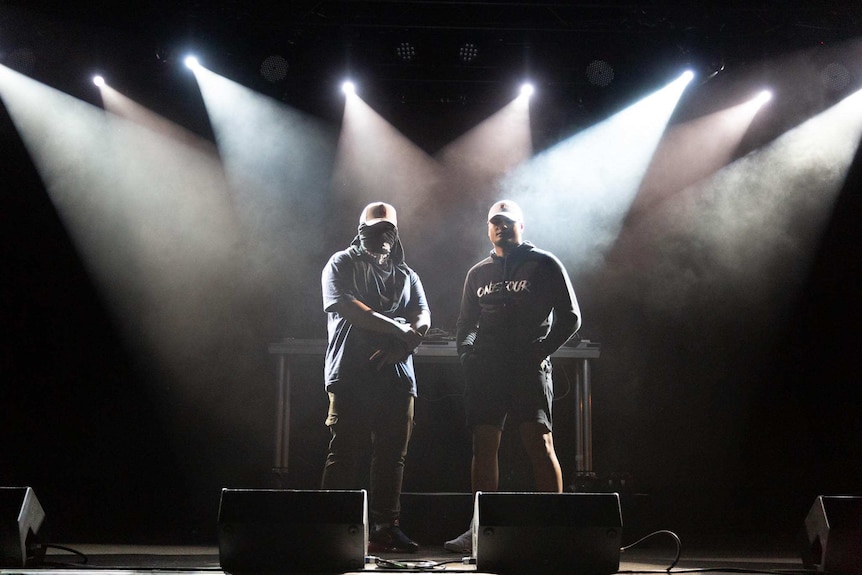 Two men stand on stage in darkness with lights behind them, one is obscuring his face with a t-shirt