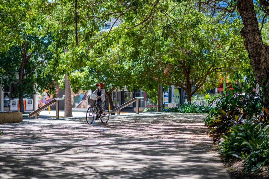 A masked woman rides her bike through Raintree Park in Darwin CBD. There is lots of greenery around her.