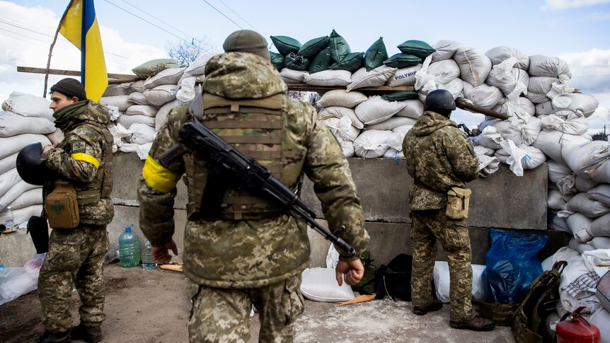 Ukrainian service members stand behind a barricade wearing yellow arm bands.