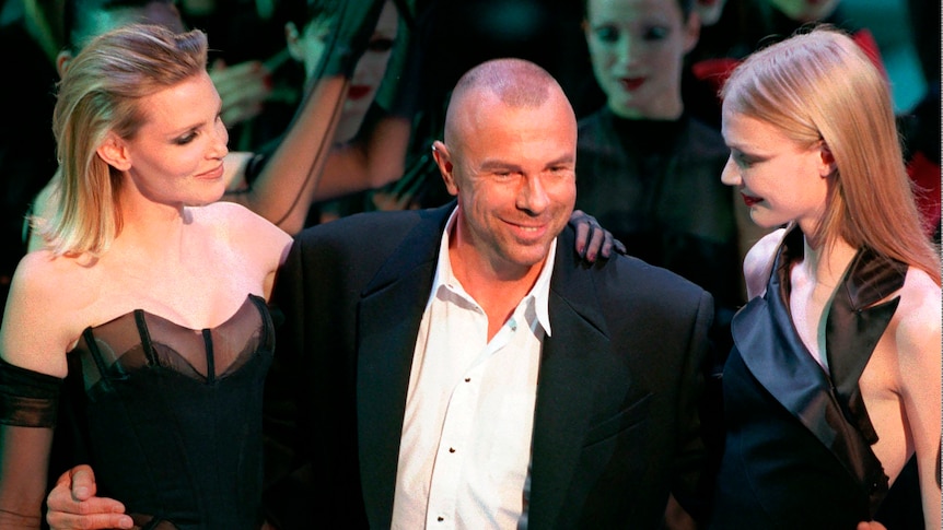 A man wearing a white shirt and black suit jacket smiles with two young women wearing black dresses either side of him.