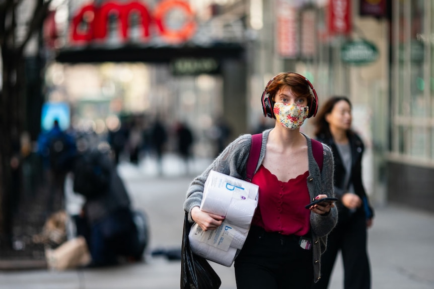 A woman walks down the street wearing headphones and a mask, carrying a phone and a pack of toilet paper.
