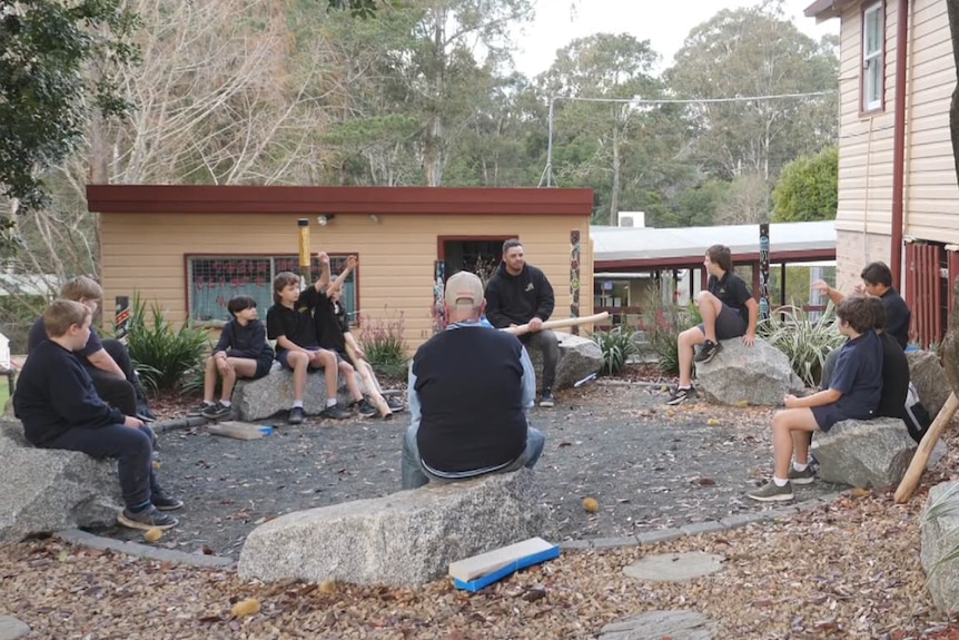 A group of young and old men sit on large rocks in a circle surrounded by buildings and landscaped gardens 