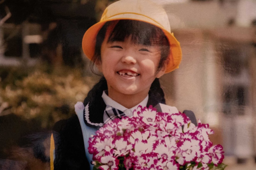 A little Japanese girl in an orange hat smiling broadly while holding a bunch of purple and white flowers