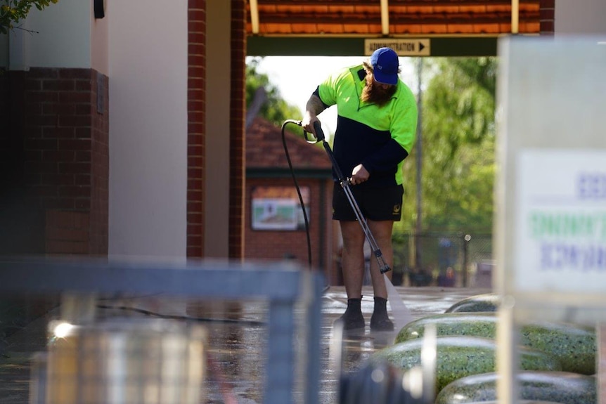 A cleaner in a hi-vis shirt and blue hat washes down a footpath with a high-pressure hose at Wembley Primary School.