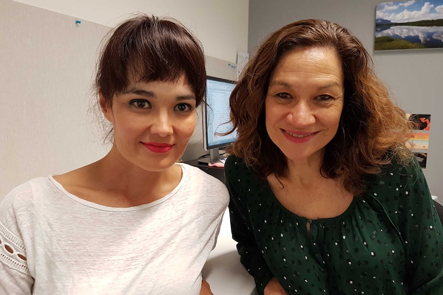 Two women smiling at the camera sitting in an office in front of a computer.