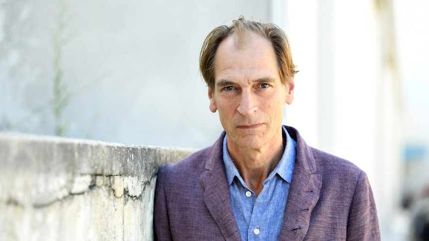 Julian Sands wearing open-neck blue shirt and purple jacket staring into camera.