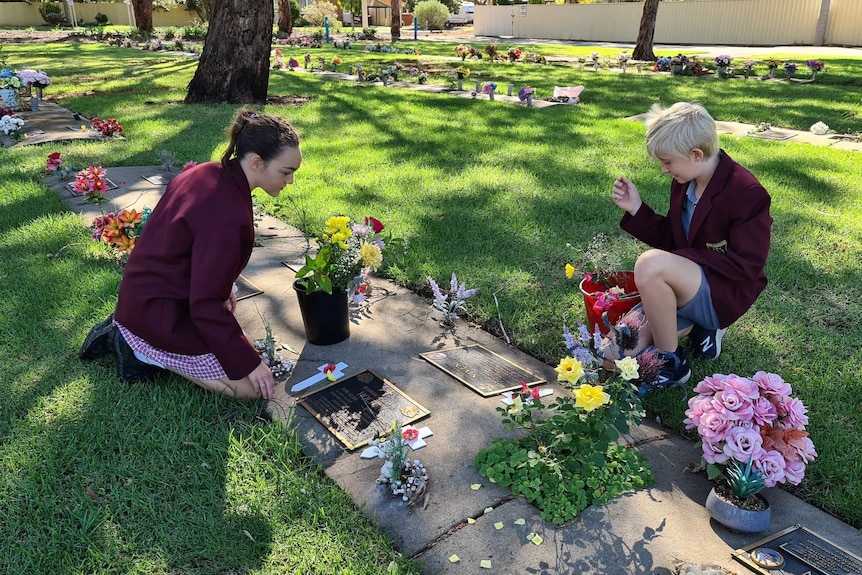 Two school students kneel down near a grave and lay flowers