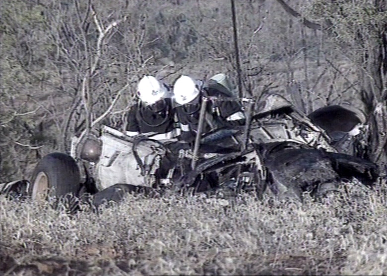 Two figures in helmets inspect mangled wreckage of helicopters