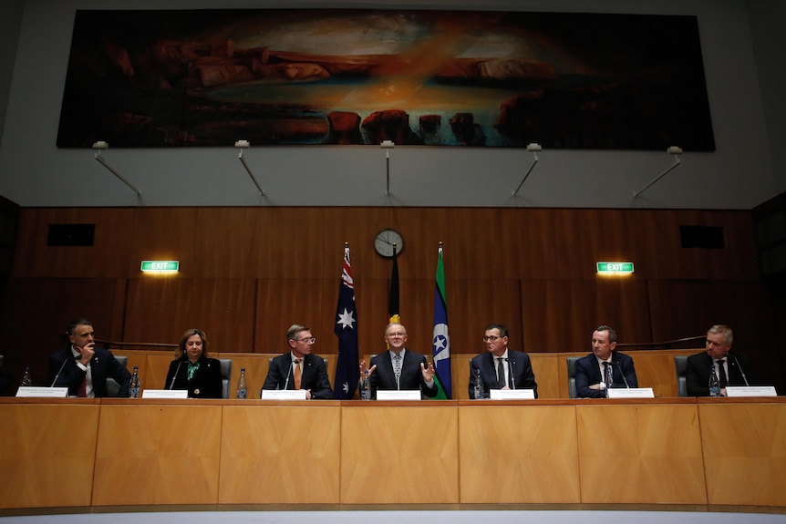 A collection of men, and one woman, sit behind a desk with microphones to address the media