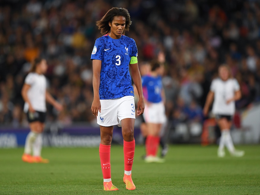 A soccer player wearing blue, white and red looks down at the ground during a game