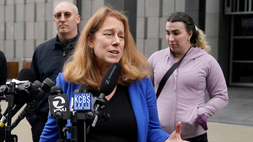 A woman in a blue suit jacket stands in front of microphones as a man and woman stand behind her. 