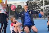 A man kneels on a netball court about to throw a pink ball, a young girl with red hair sits next to him