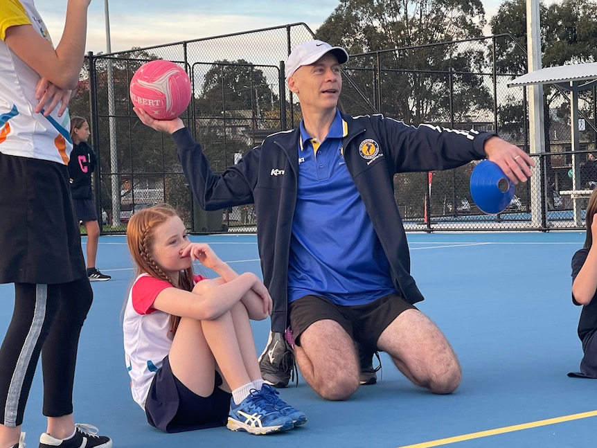 A man kneels on a netball court about to throw a pink ball, a young girl with red hair sits next to him