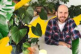 A man sits at a round dining table surrounded by his indoor plants, in a story about DIY greenhouses to help plants.