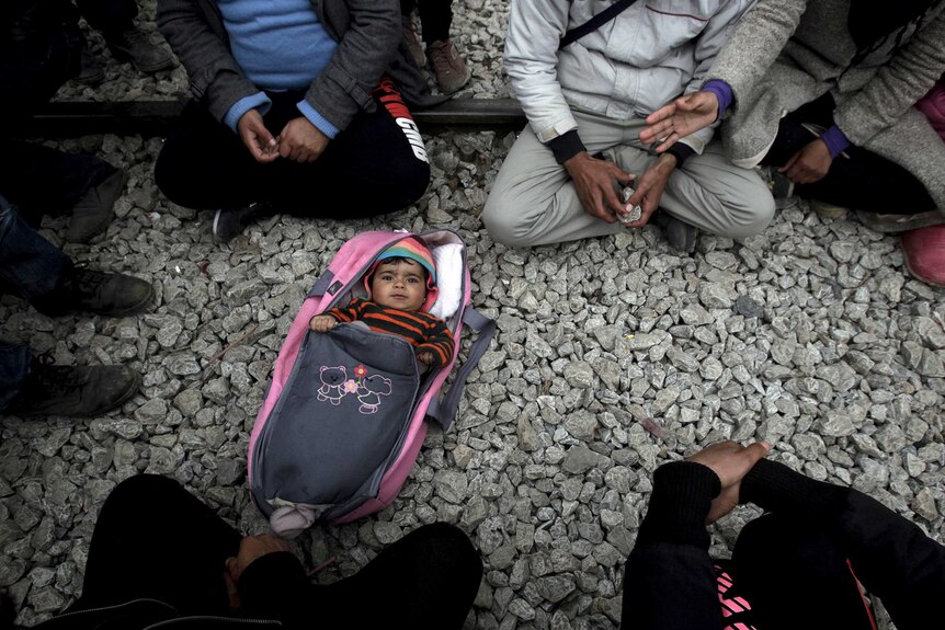 A baby lies on the ground surrounded by asylum seekers in a camp in Greece.