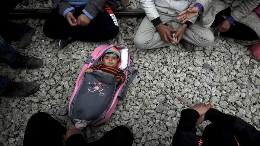 A baby lies on the ground surrounded by asylum seekers in a camp in Greece.