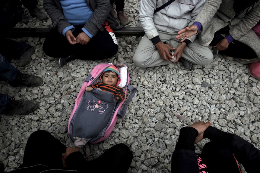 A baby lies on the ground surrounded by asylum seekers in a camp in Greece.