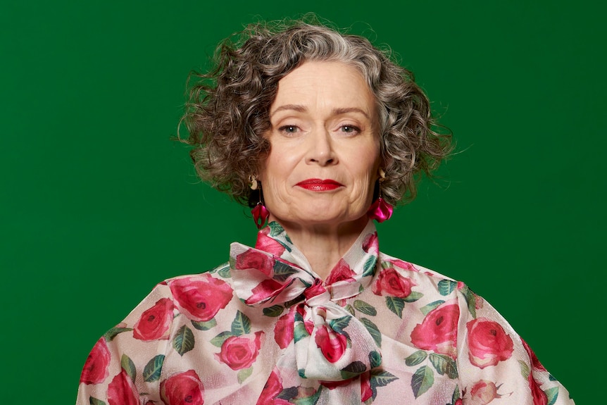 Judith Lucy, a middle-aged woman with curly, grey-streaked dark hair, looks into the lens, wearing a rose-patterned shirt.