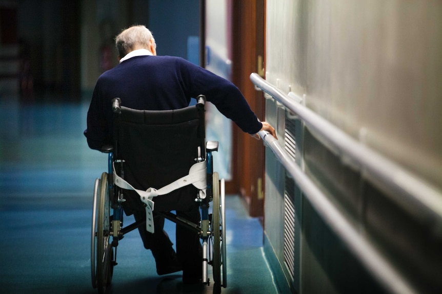 An elderly man in a wheelchair pulls himself along using handrails attached to the wall.