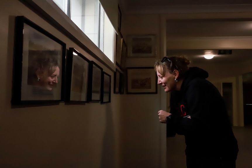 Woman bends over staring at wall of framed photos with reflection showing.