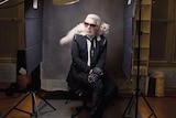 A man wearing a black suit sits on a stool in a photo studio with a white cat on his shoulders.
