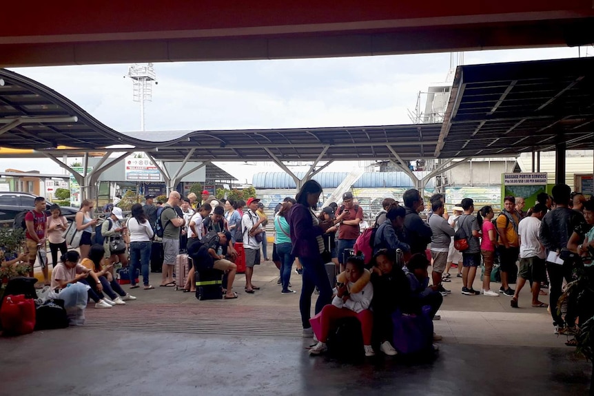 You view a crowd of travellers sitting on their bags as they under under metal shade platforms.