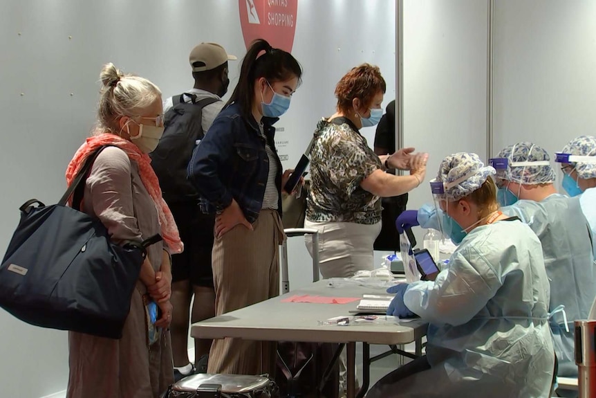 People in protective masks stand next to a bench where more people in full PPE sit.