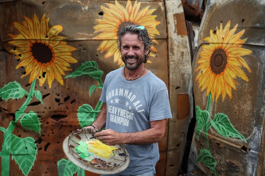 Un uomo barbuto sorridente con una camicia blu in mano davanti al lato di un'auto arrugginita ricoperta di girasoli.