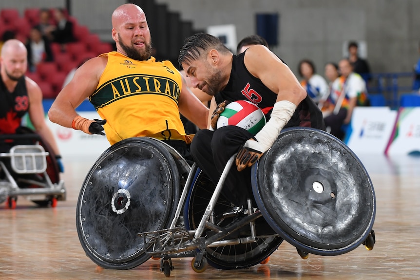 A bald Australian wheelchair rugby player collides with the wheelchair of the Canadian player holding the ball during a game.
