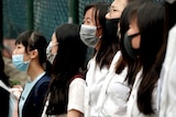 A couple of schoolgirls wear black masks, standing shoulder by shoulder. 