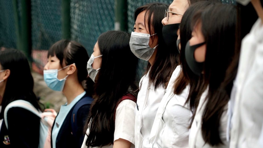 Girls in face masks are lined up shoulder to shoulder at a chain fence.