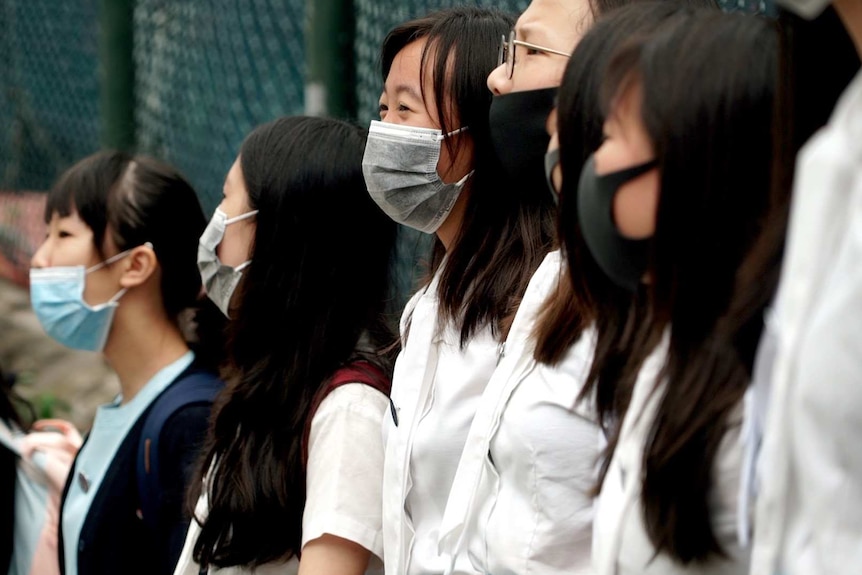 Girls in face masks are lined up shoulder to shoulder at a chain fence.