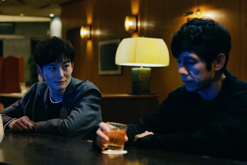 Two young Japanese men in dark clothes sit drinking whiskey in a warmly lit bar.