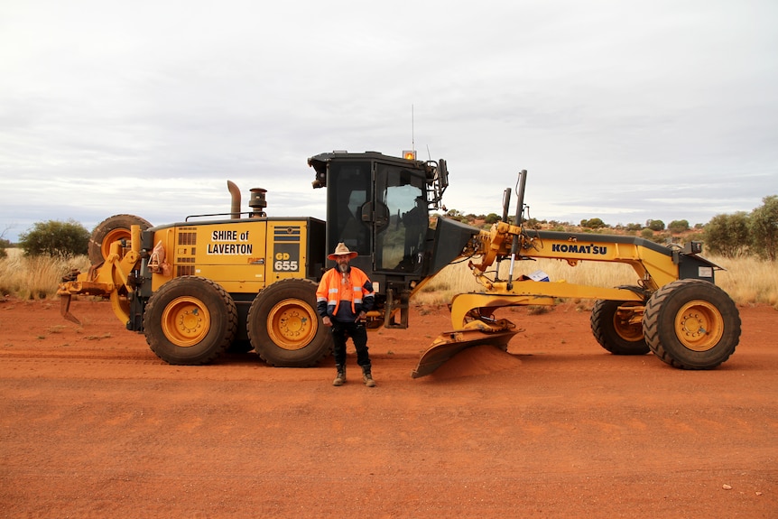 A man wearing high vis and a hat, standing in front of a road grader on a red dirt highway. 