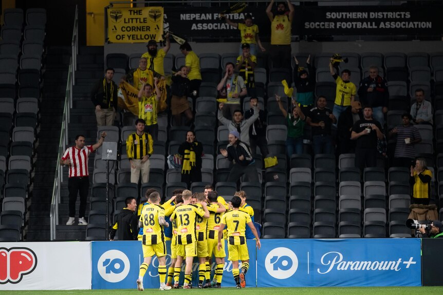 A soccer team wearing yellow and black huddles in a circle in front of fans