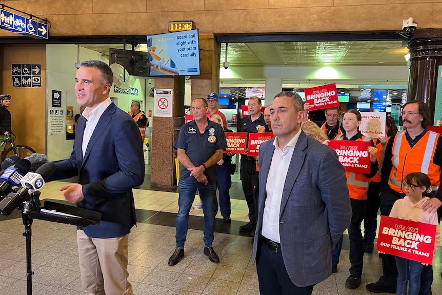 Two men in suits stand apart in a train station, behind them people are holding red signs