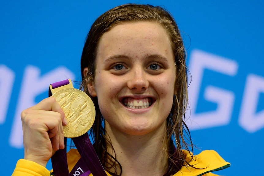 Swimmer Ellie Cole holds a Paralympic gold medal next to her face and smiles