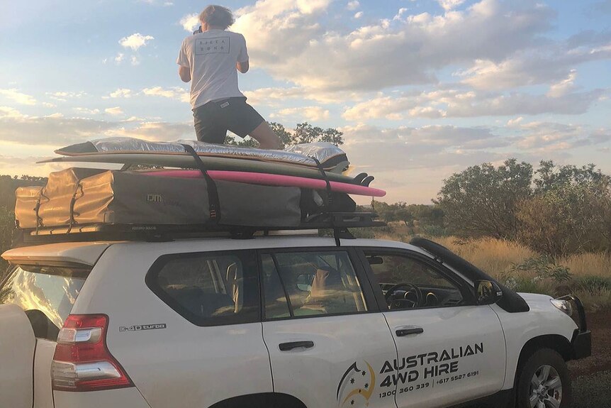 A young man faces away while using a camera and kneeling on top of a 4WD in bushland with Australian 4WD Hire logo on vehicle.