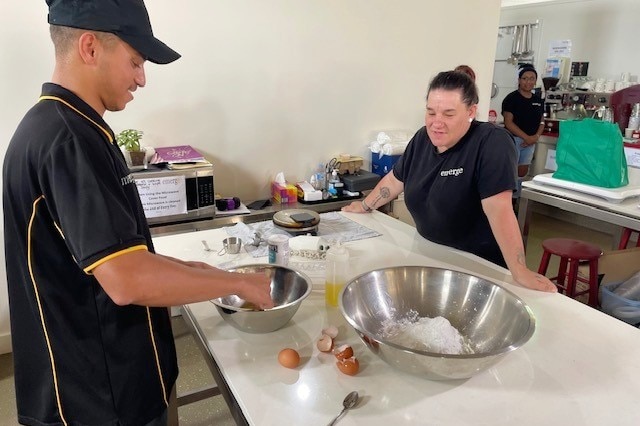 A boy cracks eggs while speaking to a woman in a kitchen.