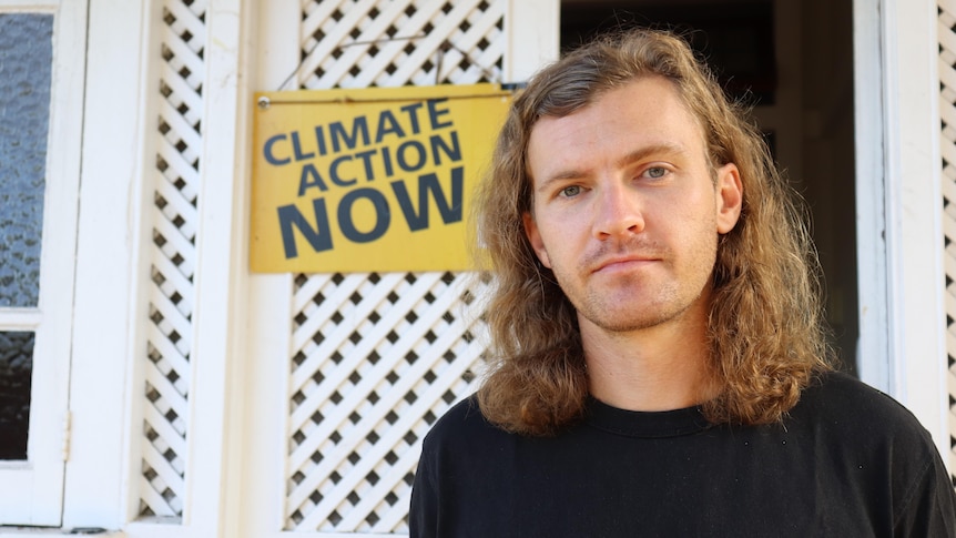 A long-haired man stands outside a house with a 'climate action now' sign behind him