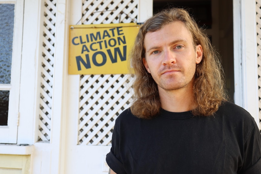 A long-haired man stands outside a house with a 'climate action now' sign behind him