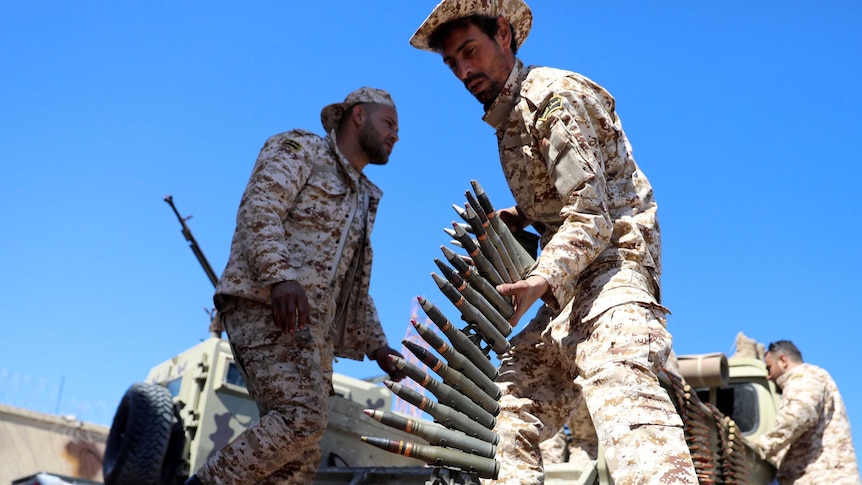 A soldier lifts a magazine of bullets. In the background are other soldiers, a military vehicle and a bright blue sky.