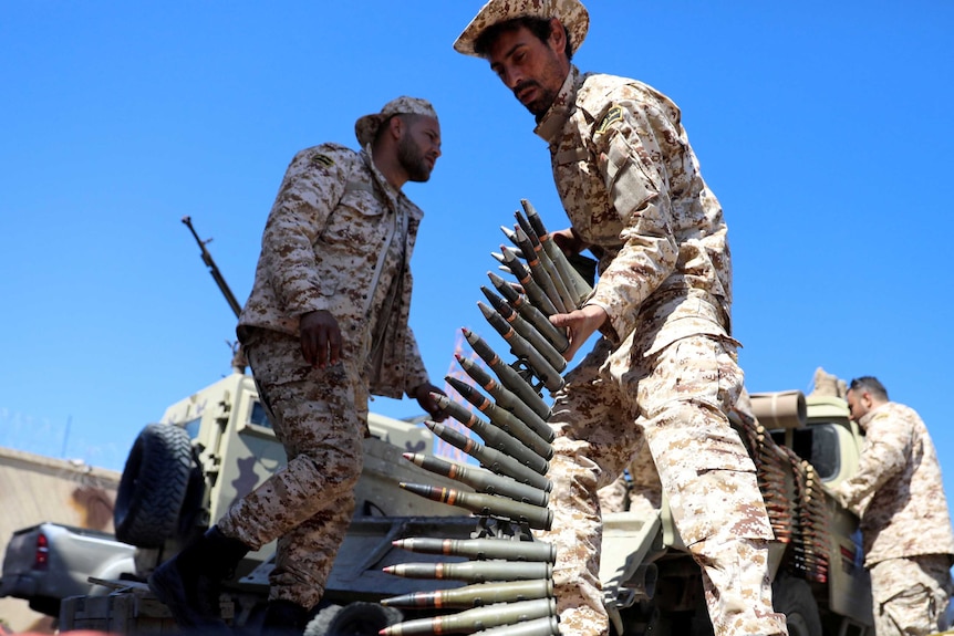 A soldier lifts a magazine of bullets. In the background are other soldiers, a military vehicle and a bright blue sky.