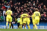 A group of Premier League footballers huddle together in celebration of a goal, while their fans roar in the background.