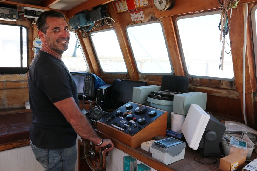 Man smiling at camera, standing at the helm of the boat in wooden cabin, instruments on dash