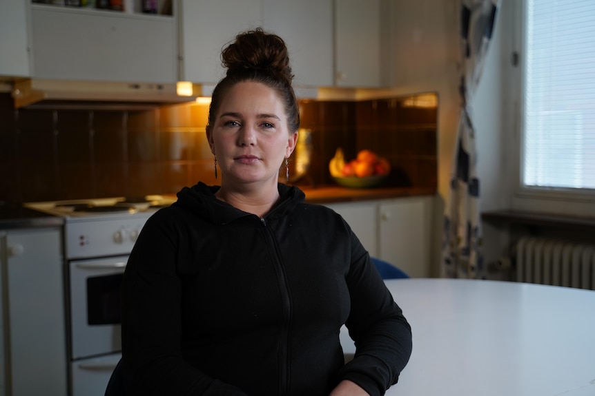 A young woman sits in her kitchen and looks into camera with a serious expression.