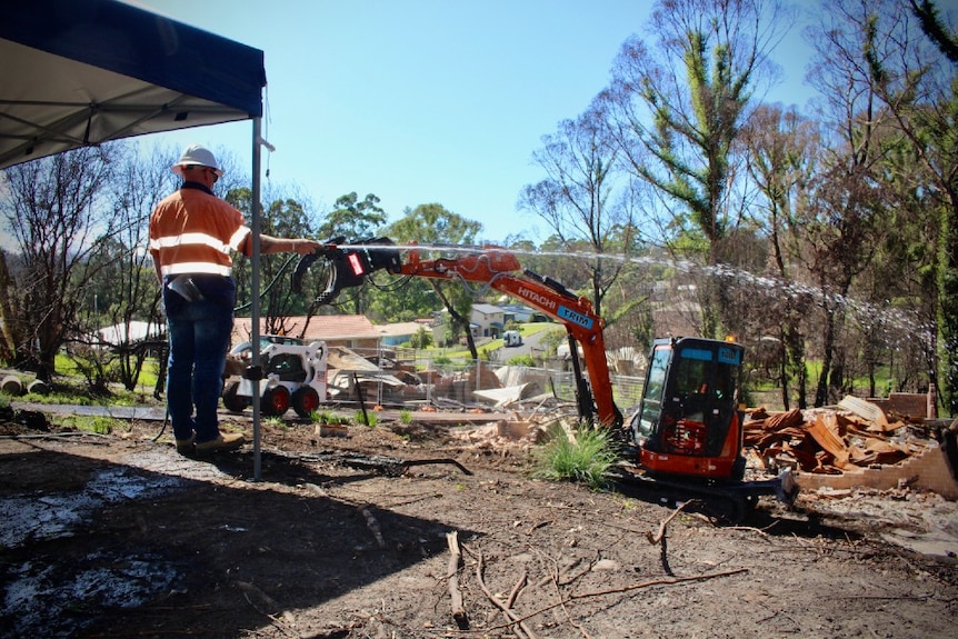 A worker in high-vis sprays a house over the ruins of a home. In the background an orange earth mover picks up debris.