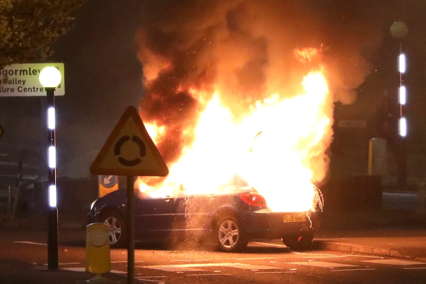 A car burns in the dark next to a roundabout road sign.
