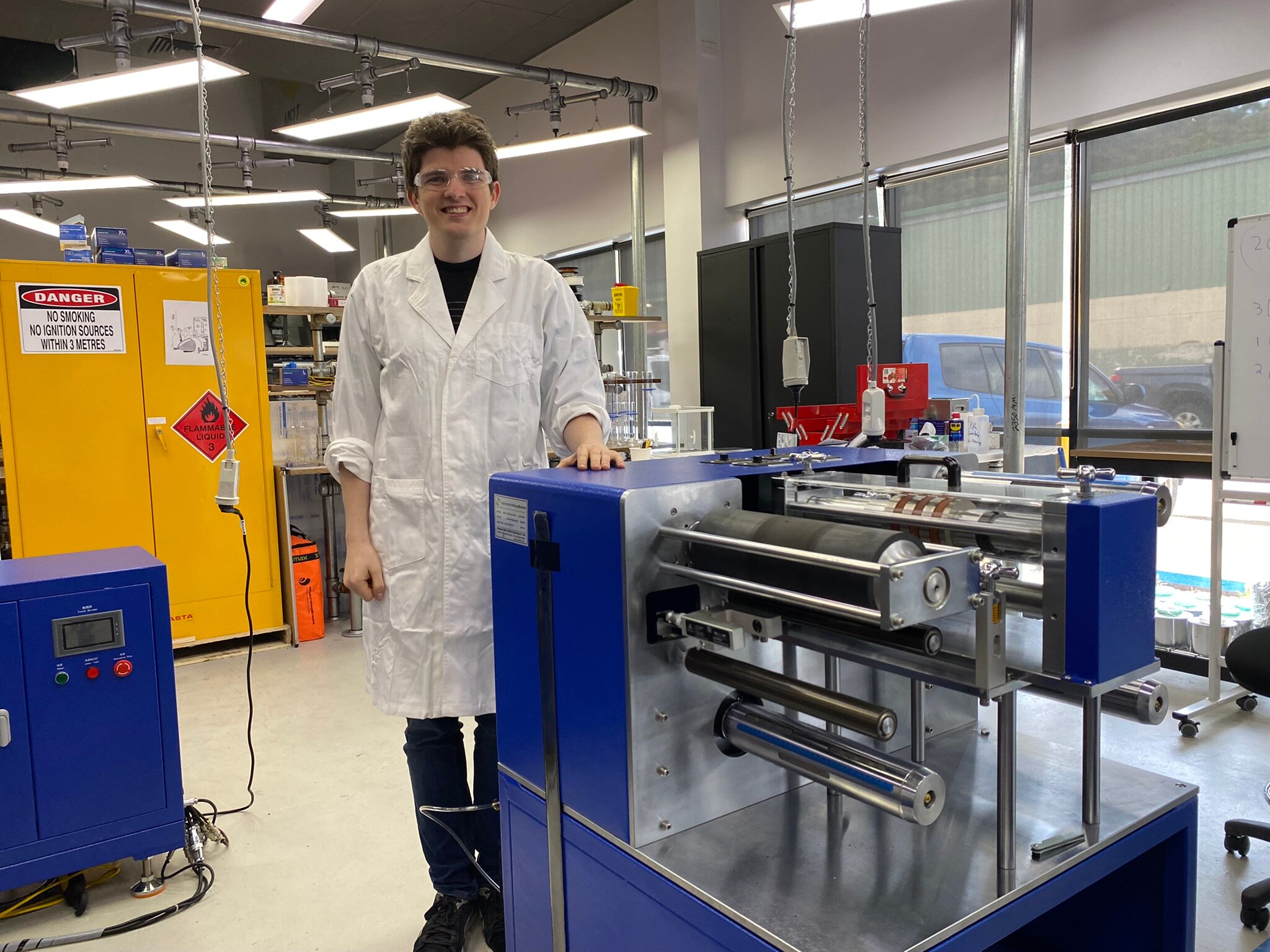 A man in a lab coat and protective glasses stands in a tech manufacturing lab.